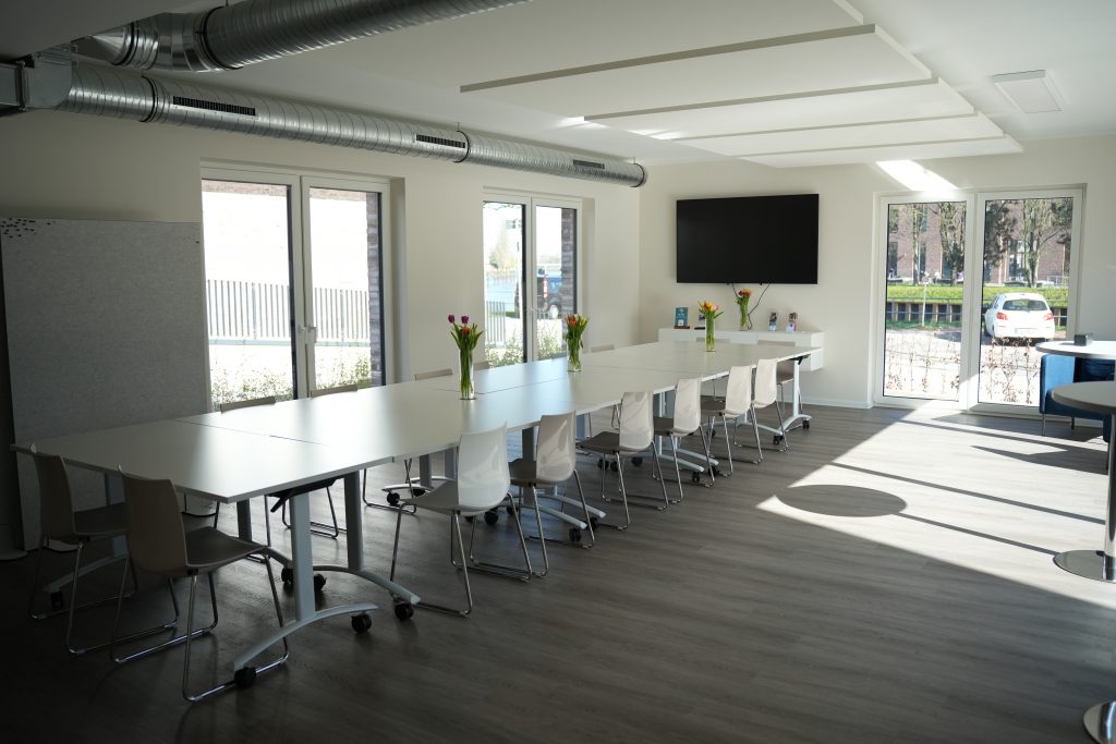 Das Bild zeigt den Seminarraum aus einem weiten Winkel. Es ist eine lange Tafel aus 8 einzelnen Tischen aufgebaut. An der Tafel stehen Stühle . Im Hintergrund ist der TV-Bildschirm zu erkennen. Die großen Fenster geben dem Raum eine helle und freundliche Atmosphäre.